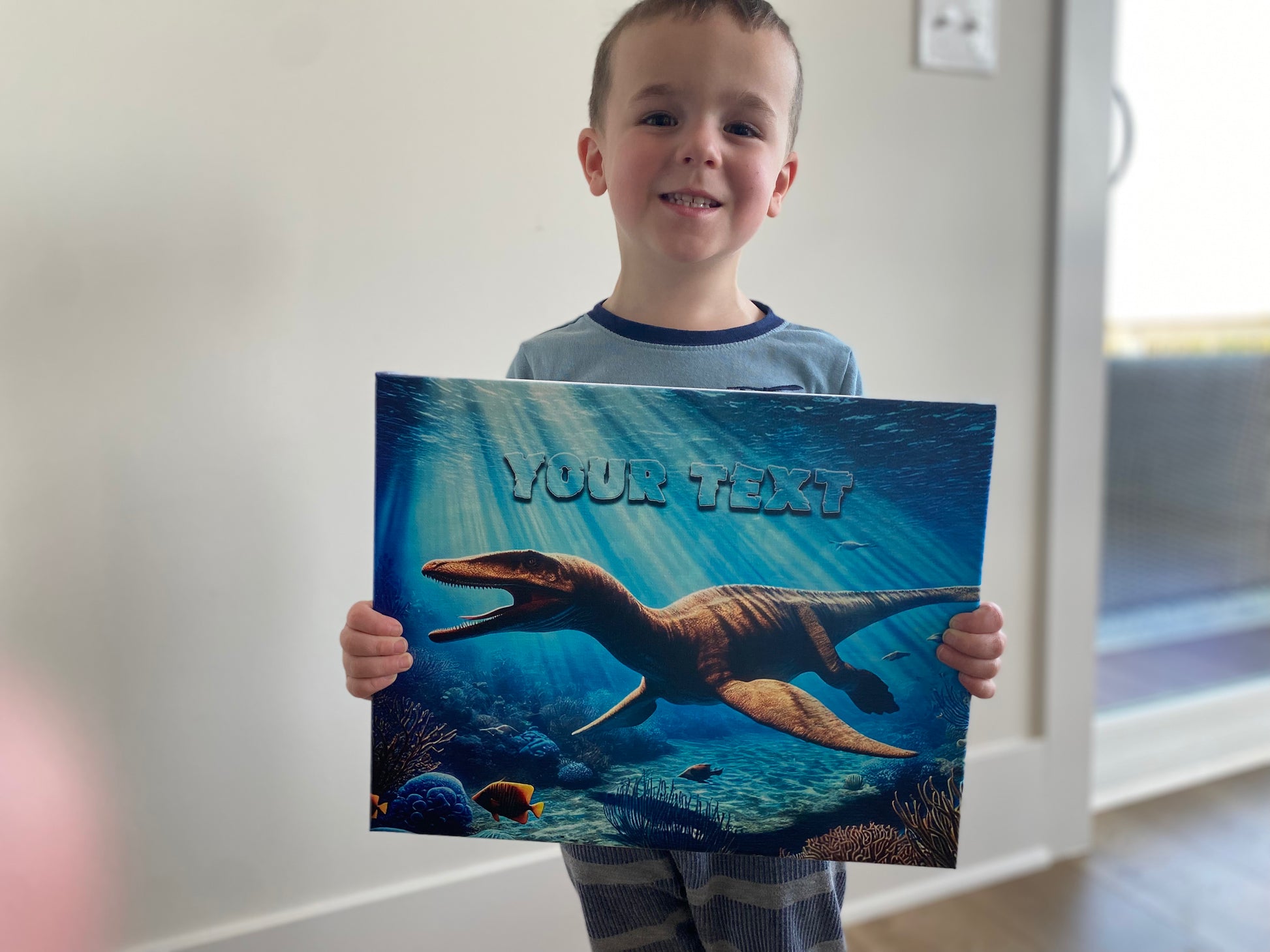 A little boy smiling holding a picture of the underwater dinosaur sign.  The sign covers the torso of his body.  Background is a white wall.
