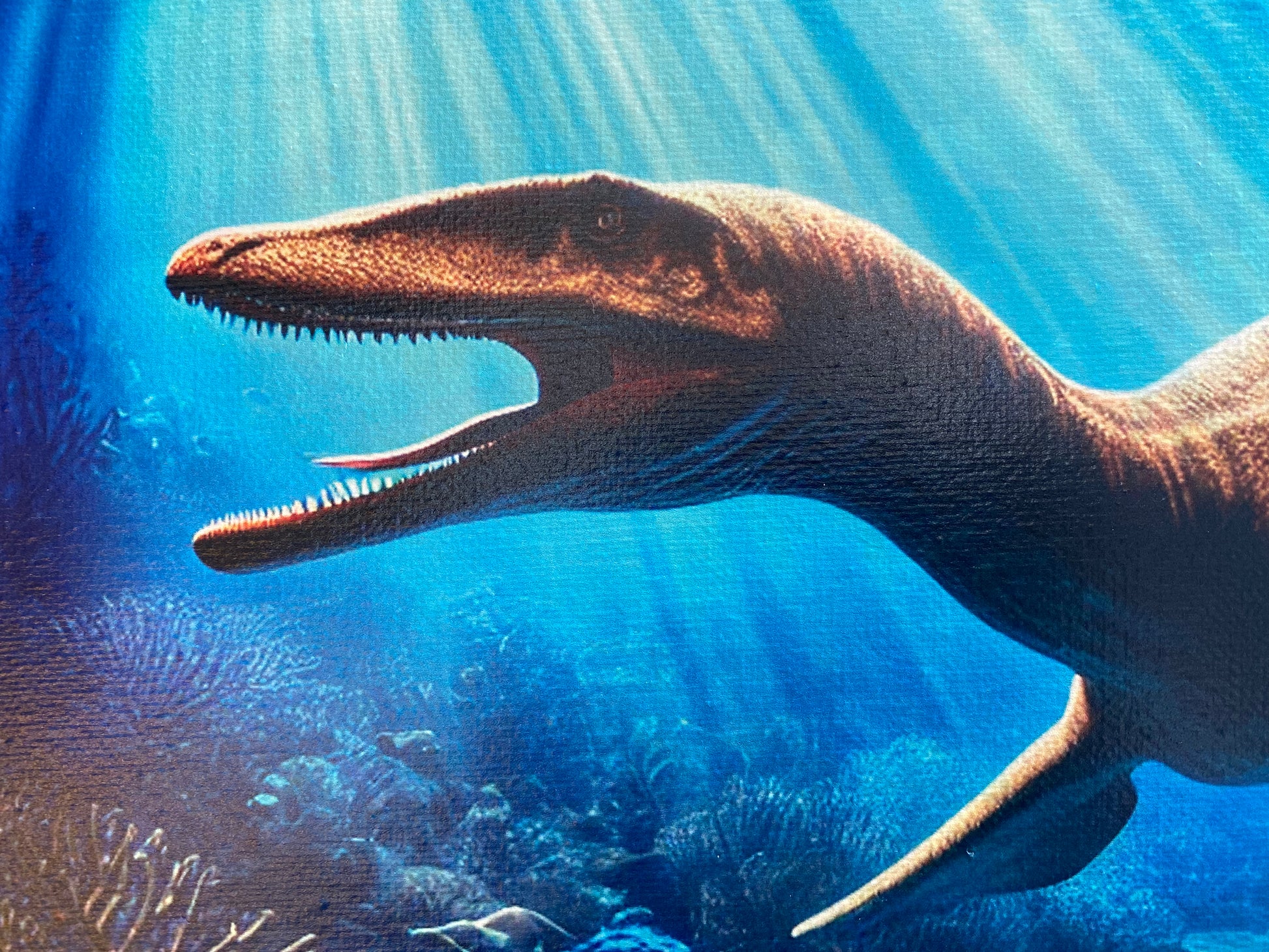 Close-up of a plesiosaur with its mouth open, revealing a tongue and tiny sharp teeth. The prehistoric marine reptile is set against a vibrant blue underwater background.