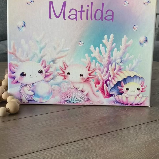 Personalized pastel canvas featuring the name "Matilda" in a playful purple font, surrounded by an enchanting underwater scene. Three adorable, pink axolotls with expressive eyes are depicted among soft-hued coral, shimmering bubbles, and a pearl-filled seashell. The dreamy watercolor-style artwork creates a whimsical oceanic theme, perfect for kids' room decor, nursery art, or a unique custom gift.