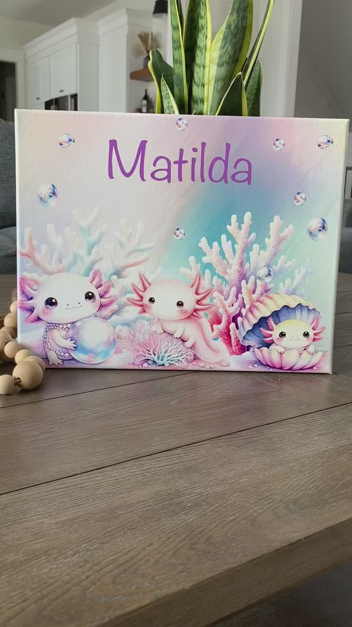 Personalized pastel canvas featuring the name "Matilda" in a playful purple font, surrounded by an enchanting underwater scene. Three adorable, pink axolotls with expressive eyes are depicted among soft-hued coral, shimmering bubbles, and a pearl-filled seashell. The dreamy watercolor-style artwork creates a whimsical oceanic theme, perfect for kids' room decor, nursery art, or a unique custom gift.